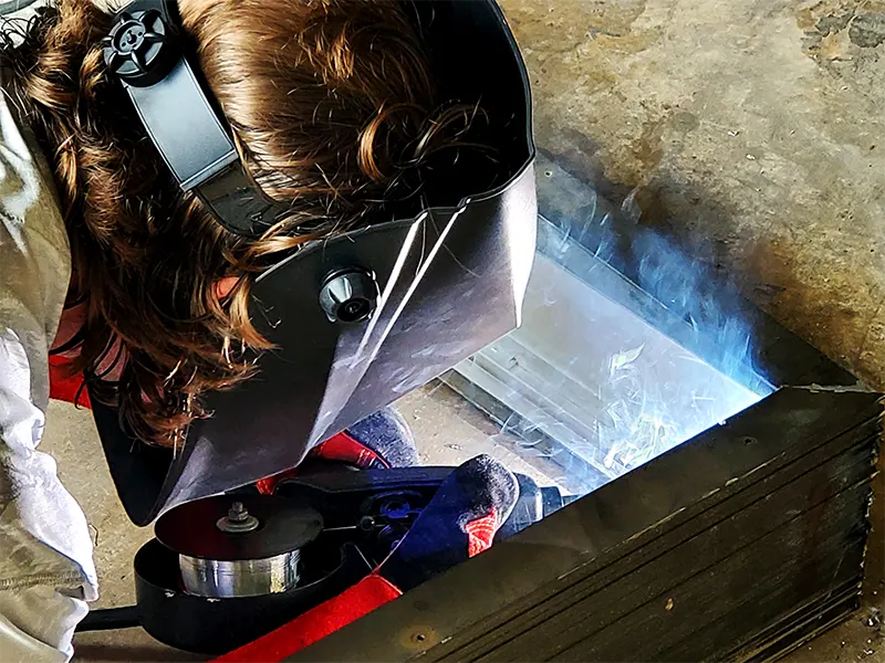 A Sparks sign fabricator in a welding helmet grinding inside a metal sign frame in the shop