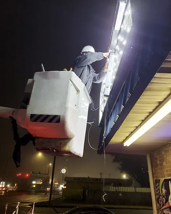 A technician in a bucket lift working on a lit storefront sign at night