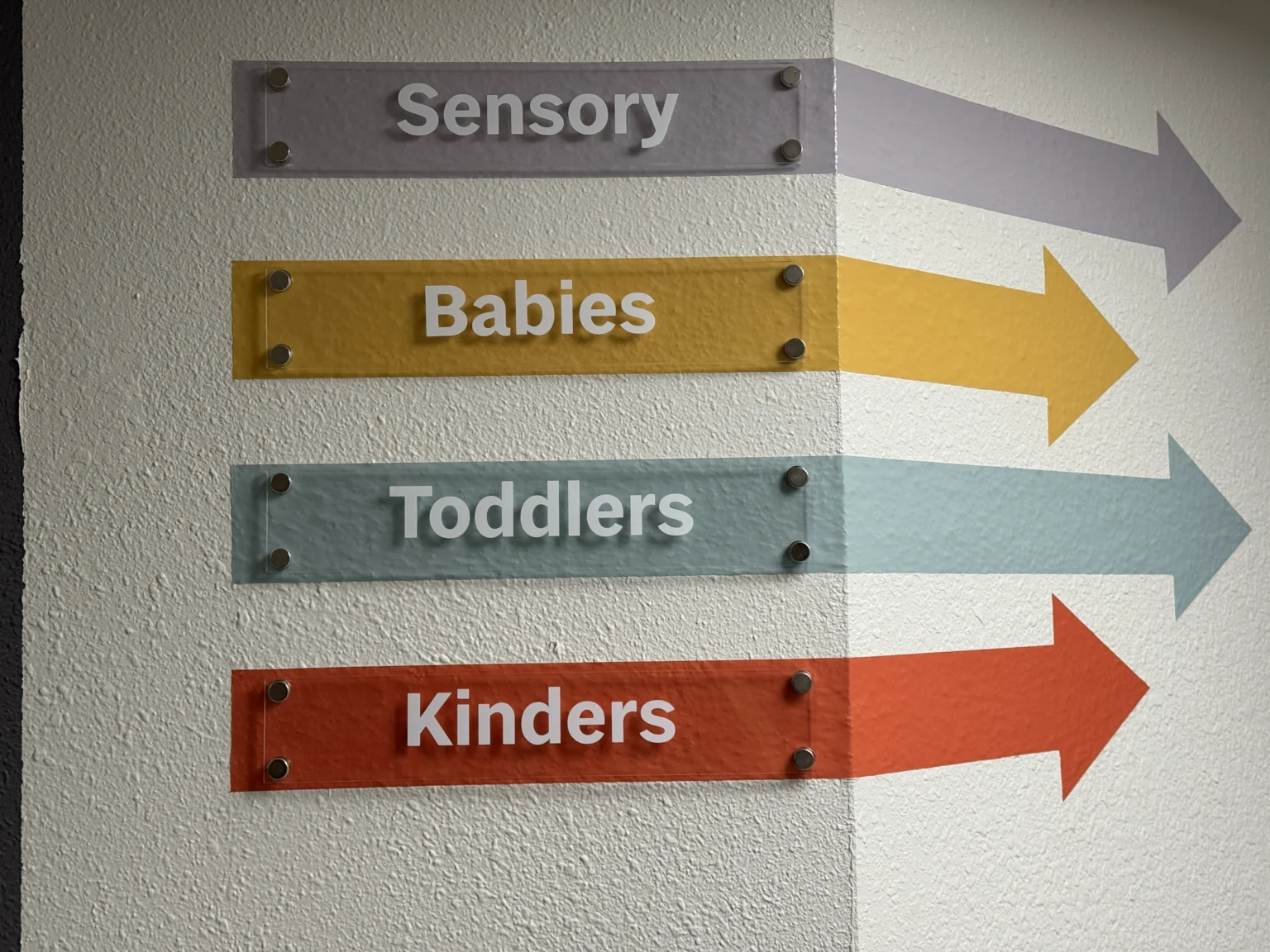 Coloured vinyl arrow signs reading Sensory, Babies, Toddlers and Kinders on a white wall at Refuge Kids