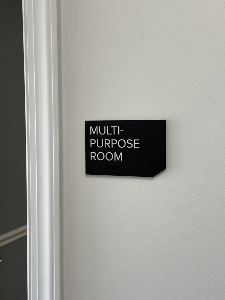 A black Multi-Purpose Room sign with a cut corner beside a white door frame, raised white letters over a row of Braille dots