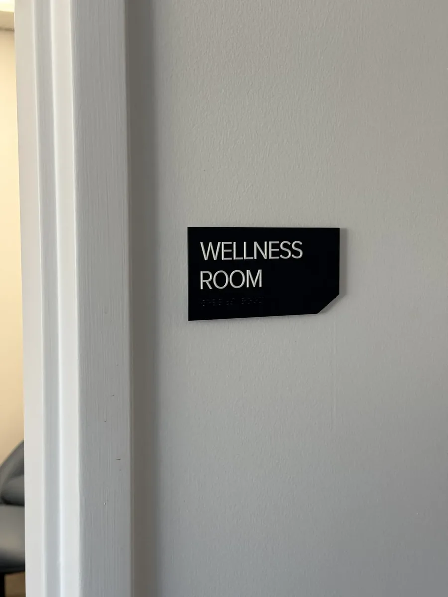 A black Wellness Room sign, one of a set of ADA braille signs, mounted beside a door frame with raised white letters and Braille dots underneath