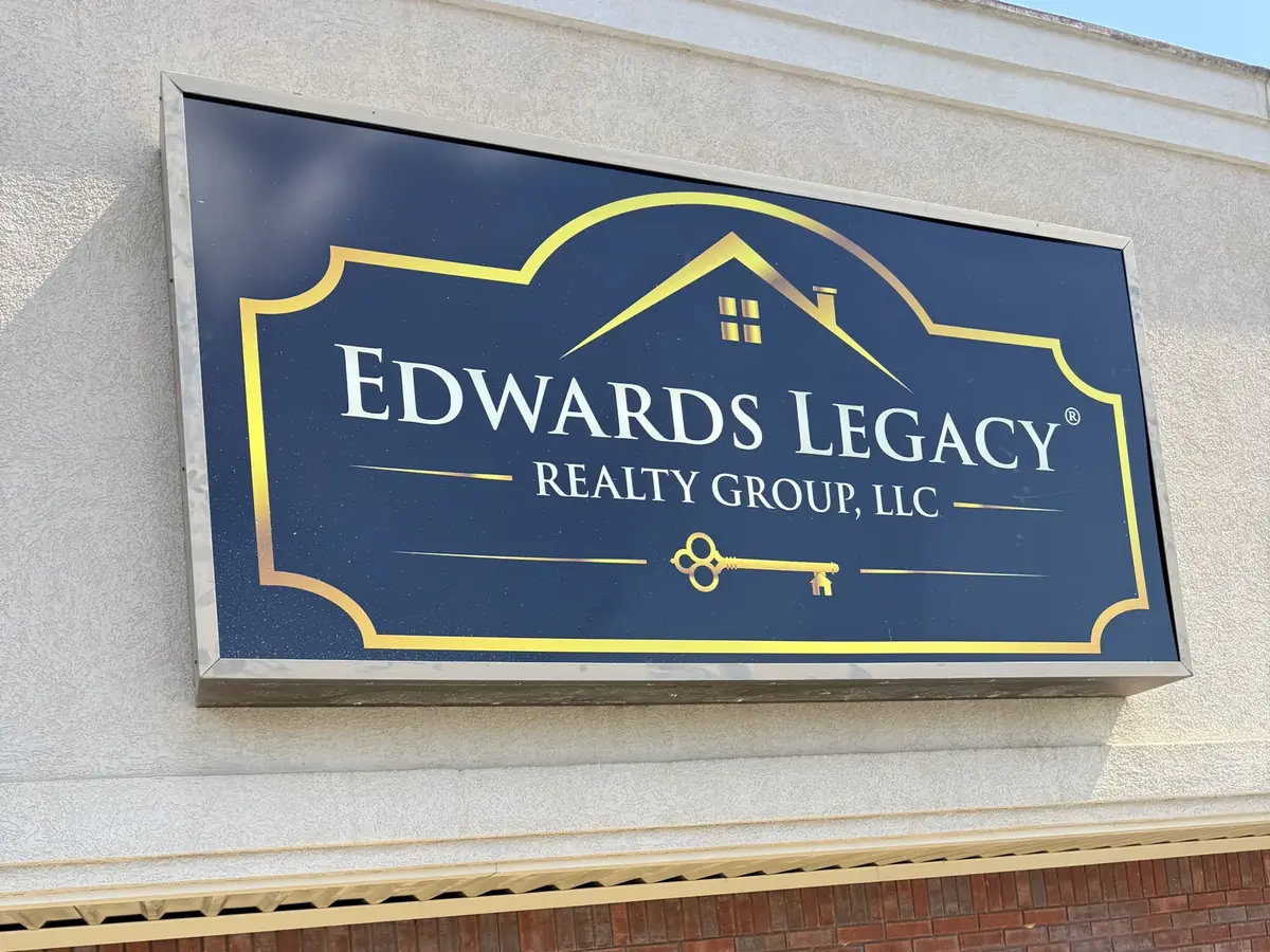 The Edwards Legacy Realty Group, LLC cabinet sign on a strip-center fascia, gold logo and lettering on a navy face above a brick storefront