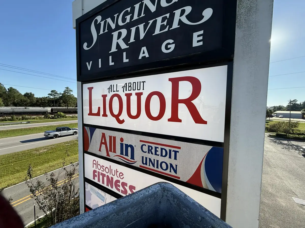 A shopping center pylon by a highway, the center's name on the top cabinet and each tenant's name on its own panel below