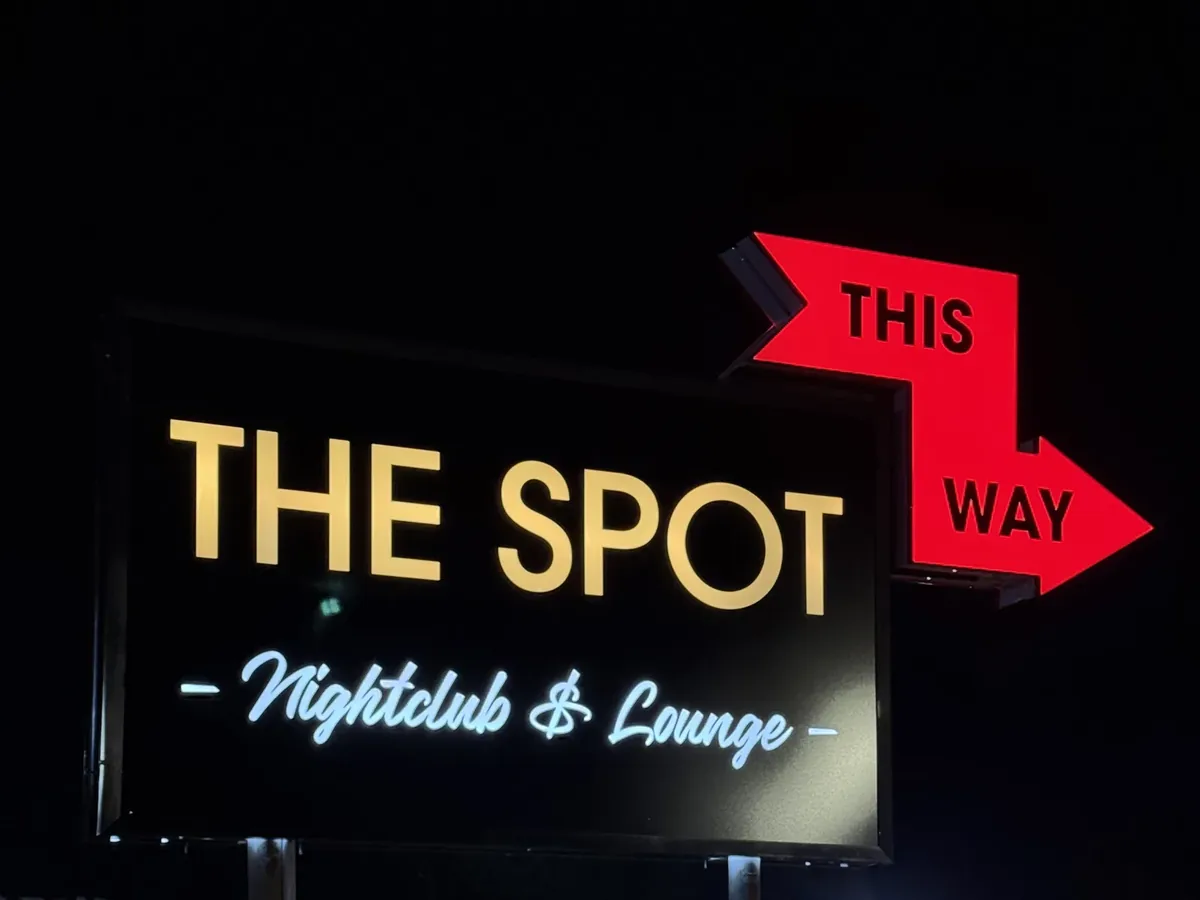 The Spot Nightclub & Lounge cabinet sign lit at night, gold letters and a white script line on its black face beside a red This Way arrow