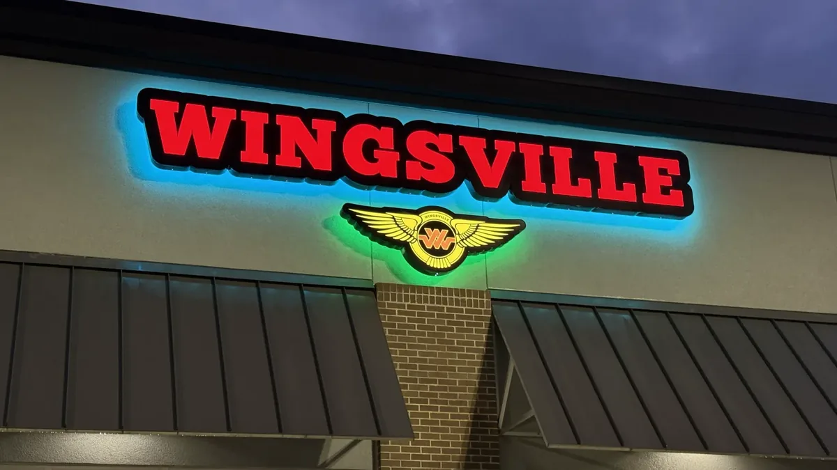 Red channel letters and a winged logo lit at dusk over a storefront, with a blue and green glow on the wall behind