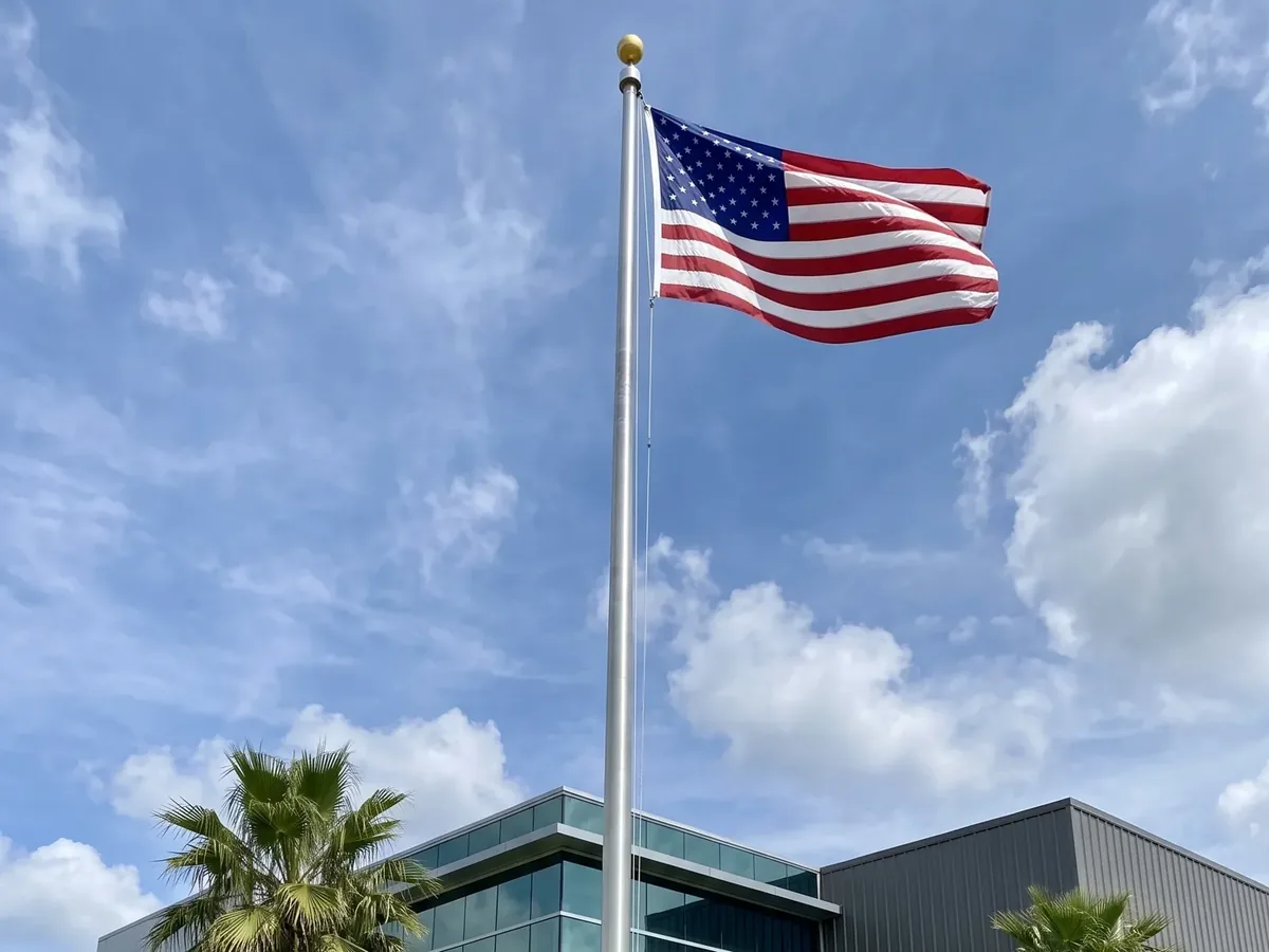 Generated illustration of a tall aluminum commercial flagpole flying a United States flag above a glass office building with palm trees