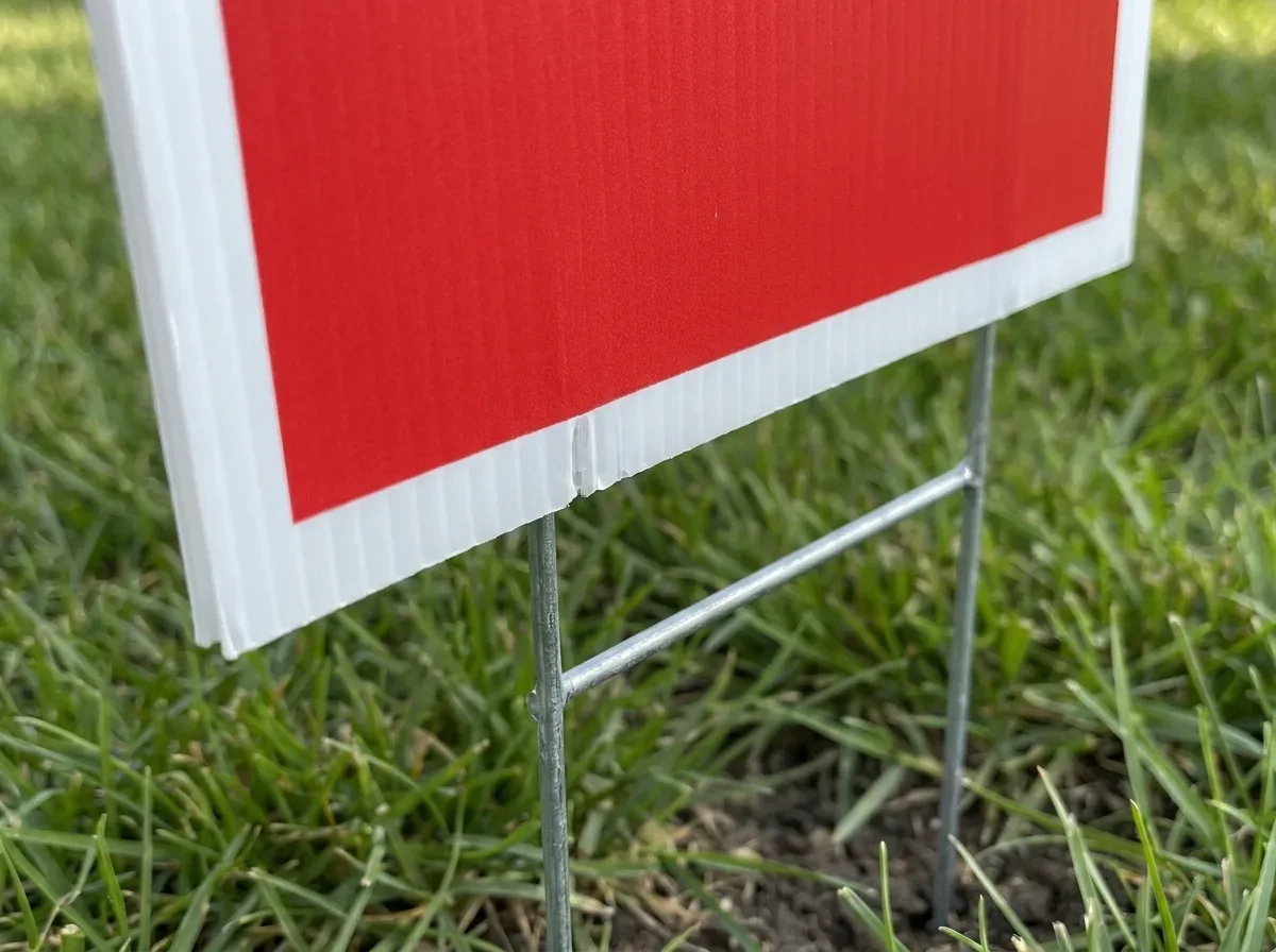 Close-up of a corrugated plastic yard sign on a galvanized H-wire stake pressed into grass, the flutes visible along its edge