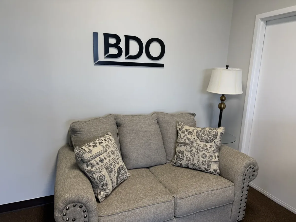 Black dimensional company logo on a lobby wall above a grey sofa and a floor lamp