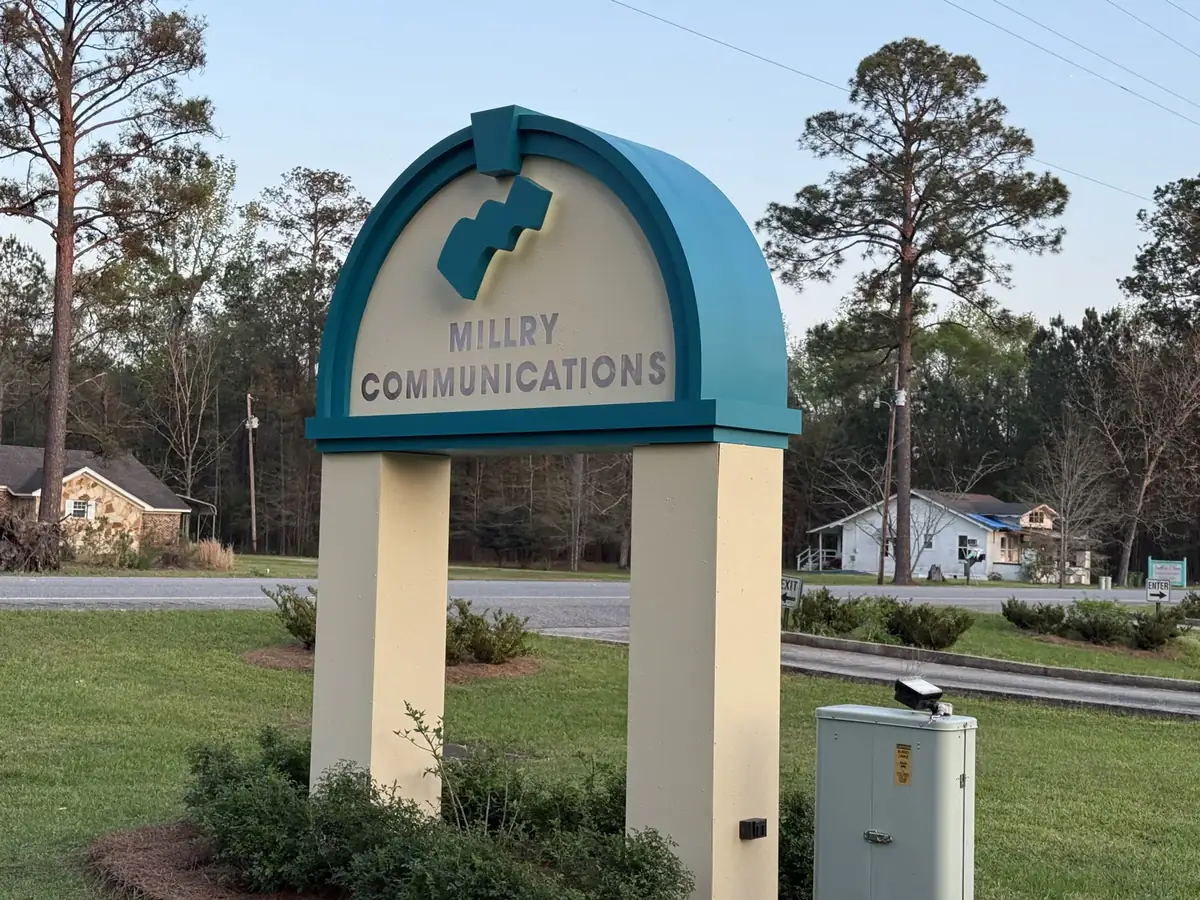 An arched monument sign with a teal frame and a cream face carrying raised dimensional letters and a logo, standing on two cream posts in a planted bed on a lawn at dusk