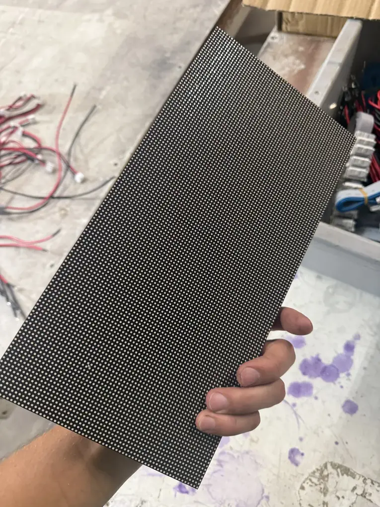 An LED display module for outdoor digital signage, held in a hand in the shop, its face covered in tiny LED pixels