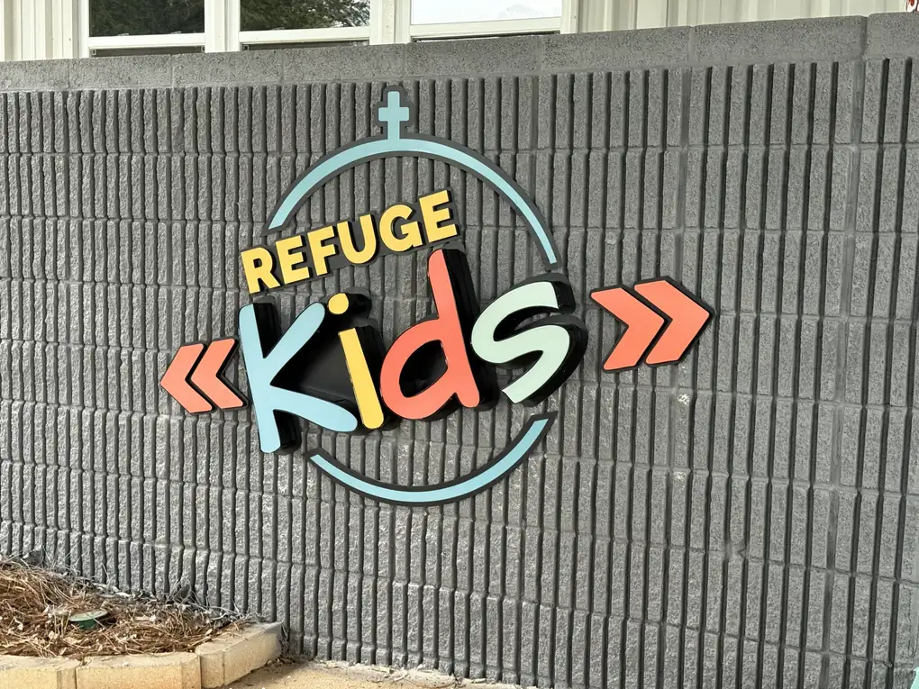 A layered Refuge Kids logo sign, a non-illuminated outdoor sign mounted on a grey block wall