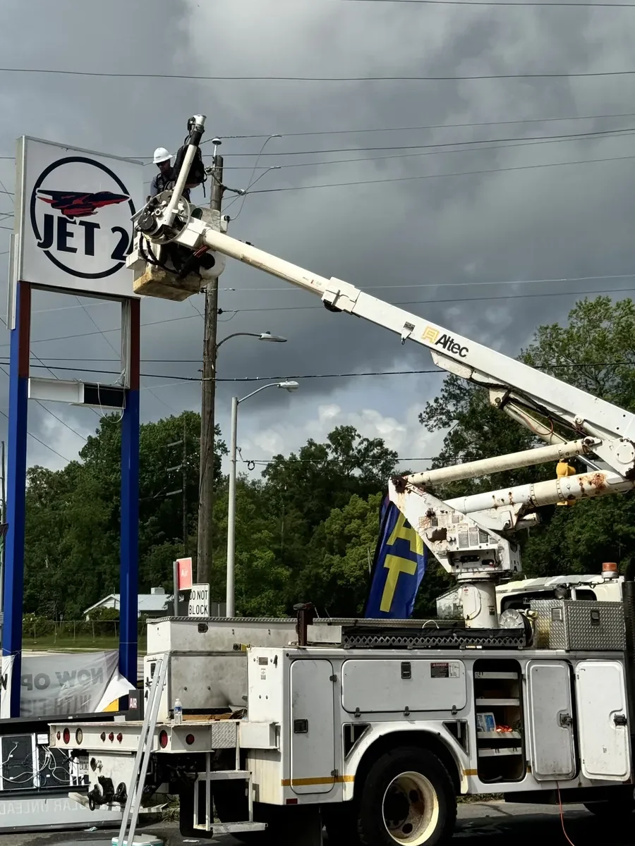 Crew member in a bucket truck's bucket at the top of a convenience store's pylon sign under storm clouds