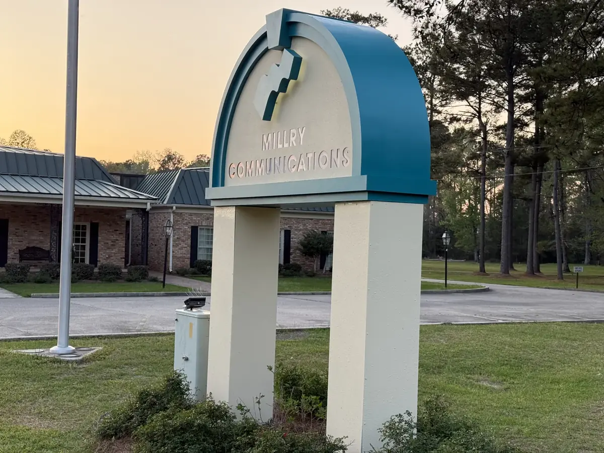 An arched teal and cream monument sign for Millry Communications at sunset, in front of a brick office building with a flagpole on the lawn