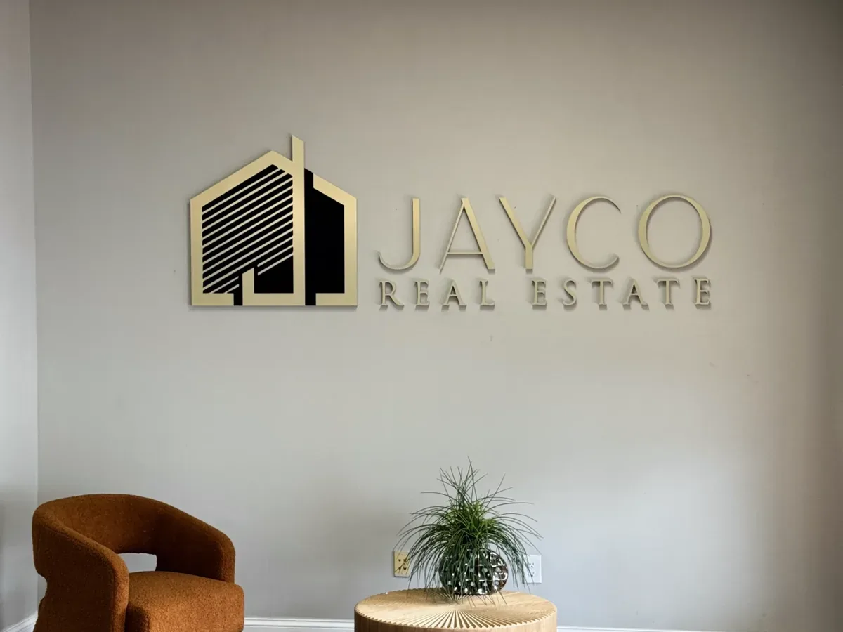 Gold and black dimensional Jayco Real Estate logo on a grey lobby wall above an armchair and a side table
