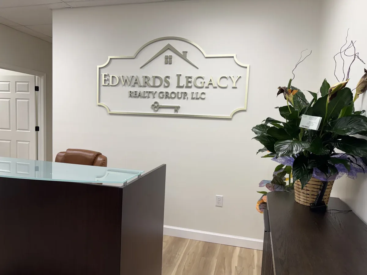 Silver dimensional Edwards Legacy Realty Group lobby sign with a house roofline and a key, on the wall above a glass-topped reception desk