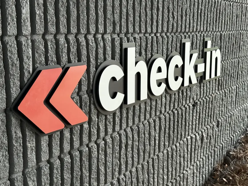 White dimensional check-in letters and two orange chevrons standing off a grey split-face block wall