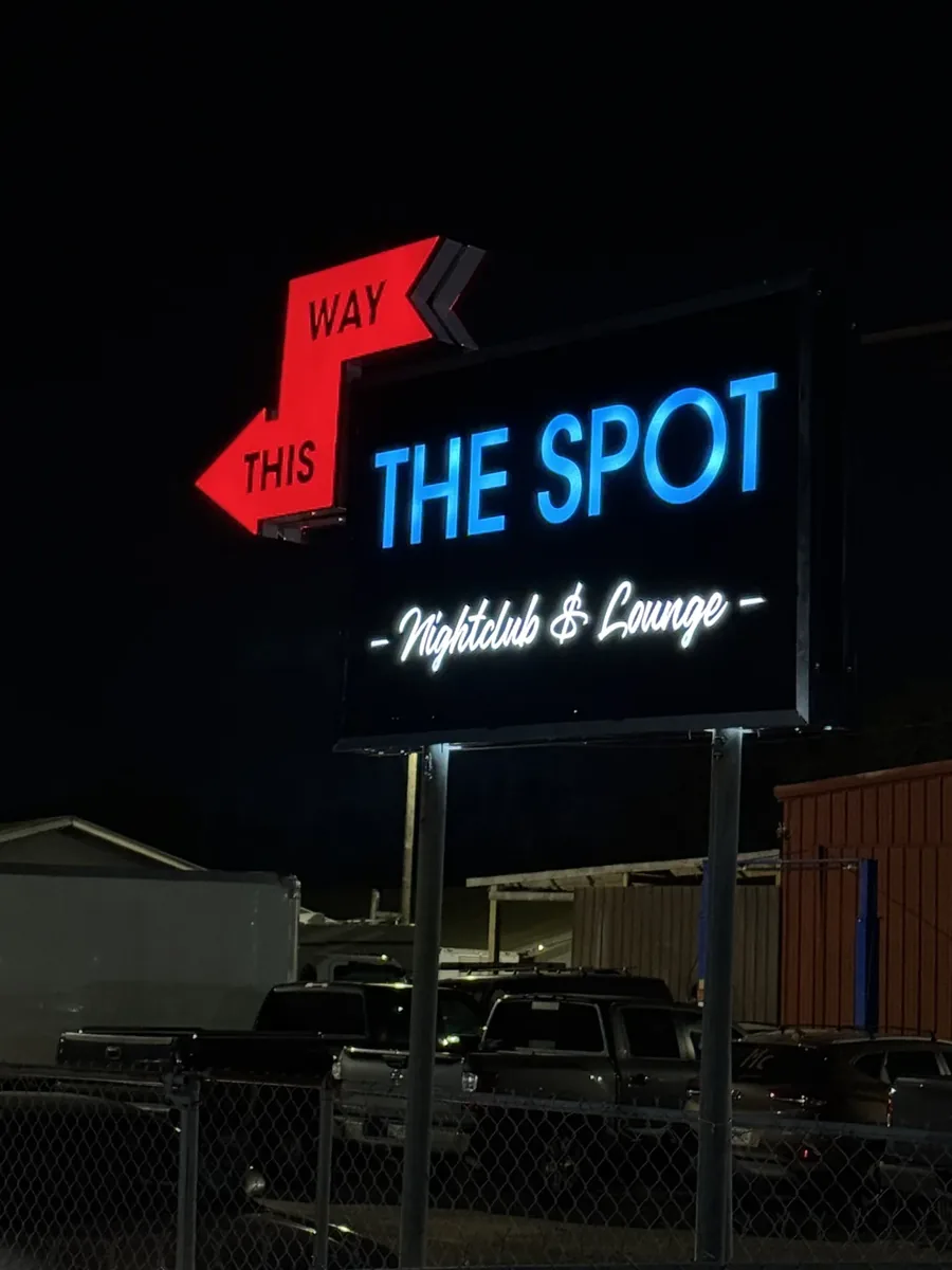The Spot Nightclub & Lounge cabinet sign lit at night, blue letters and a white script line glowing on its black face, with a red This Way arrow at its corner
