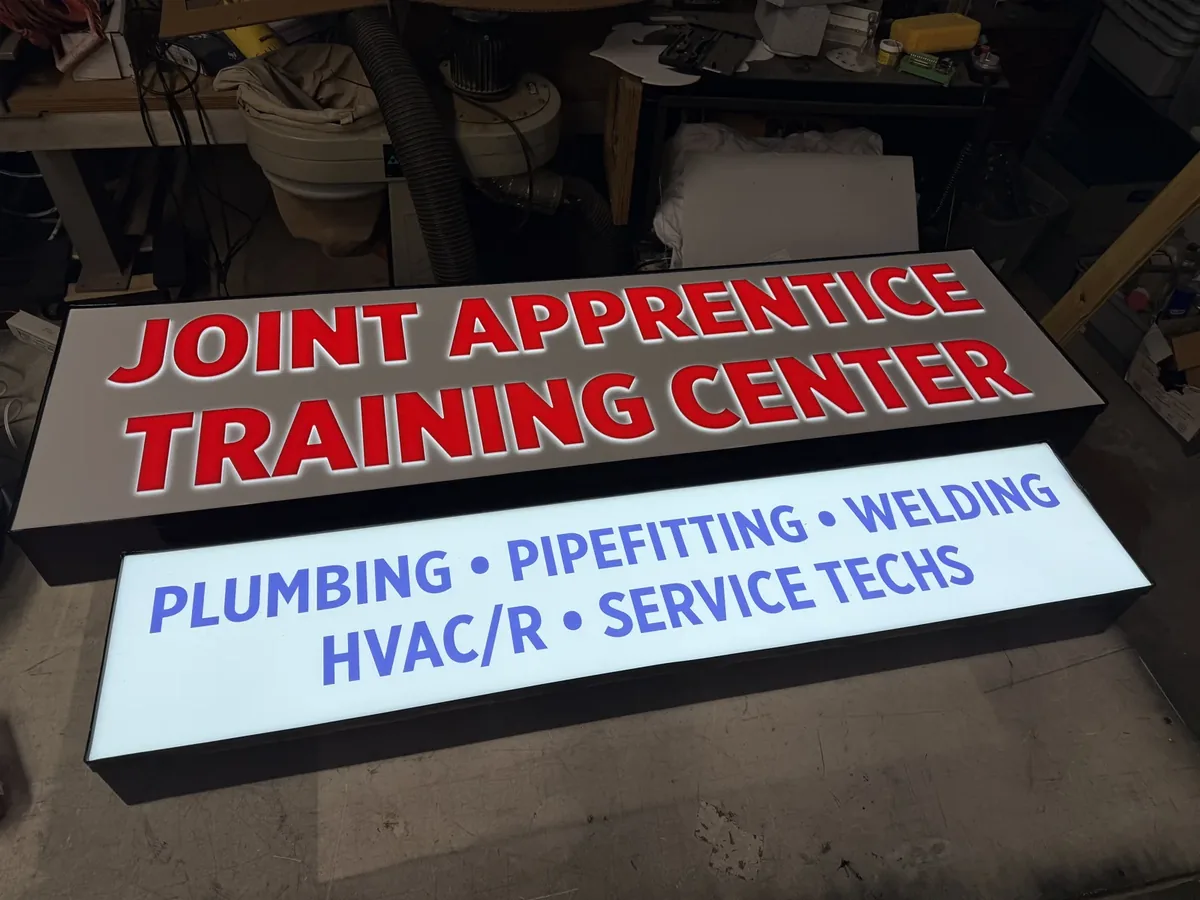 Two Joint Apprentice Training Center cabinet signs lit up on the floor of our sign shop, red letters on a grey face above a white face with blue lettering