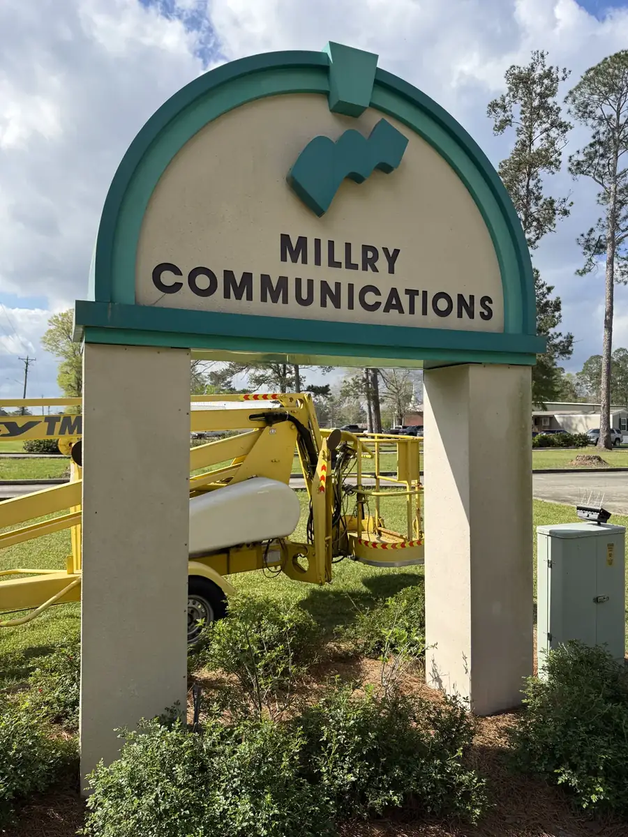 The arched teal and cream Millry Communications monument sign, with a yellow towable lift parked behind it