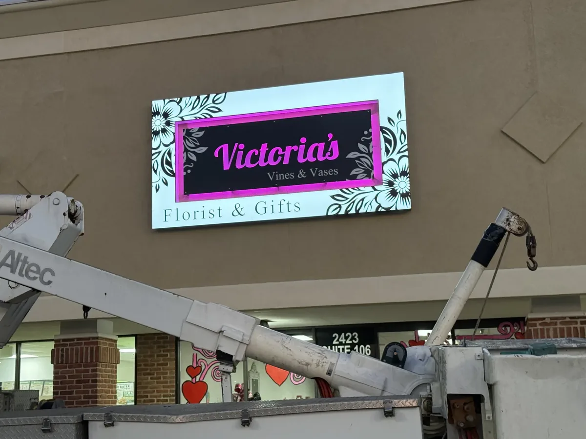 A lit florist's sign on a strip-center wall at dusk, with a bucket truck's boom in front of it