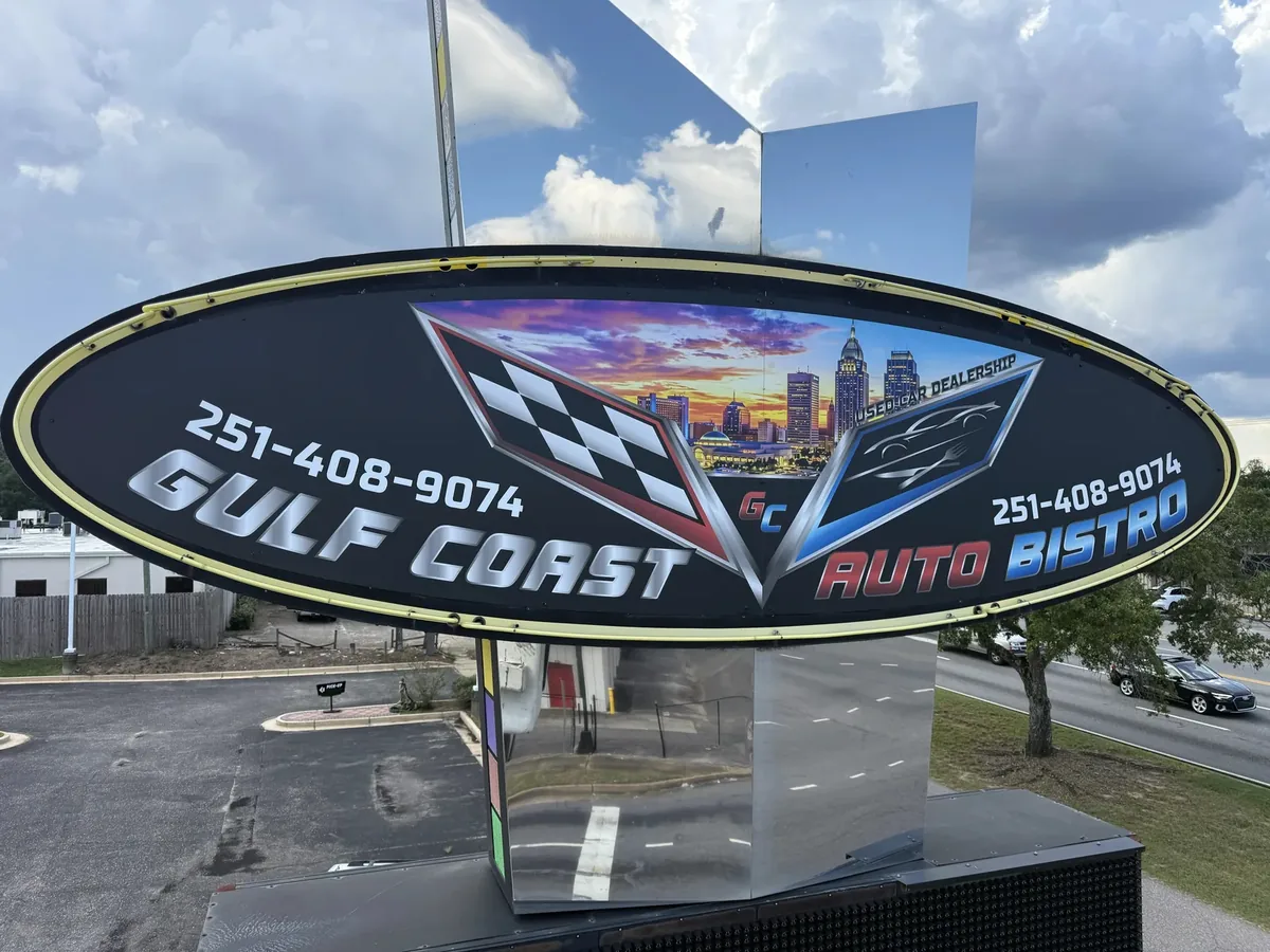 The Gulf Coast Auto Bistro oval pylon sign cabinet, a checkered-flag and city skyline graphic on a black oval, standing over a parking lot by the road
