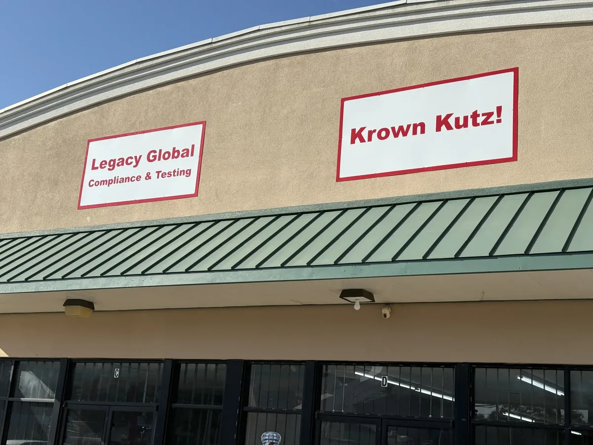 Legacy Global Compliance & Testing flat sign panel above a green awning, beside a Krown Kutz! panel