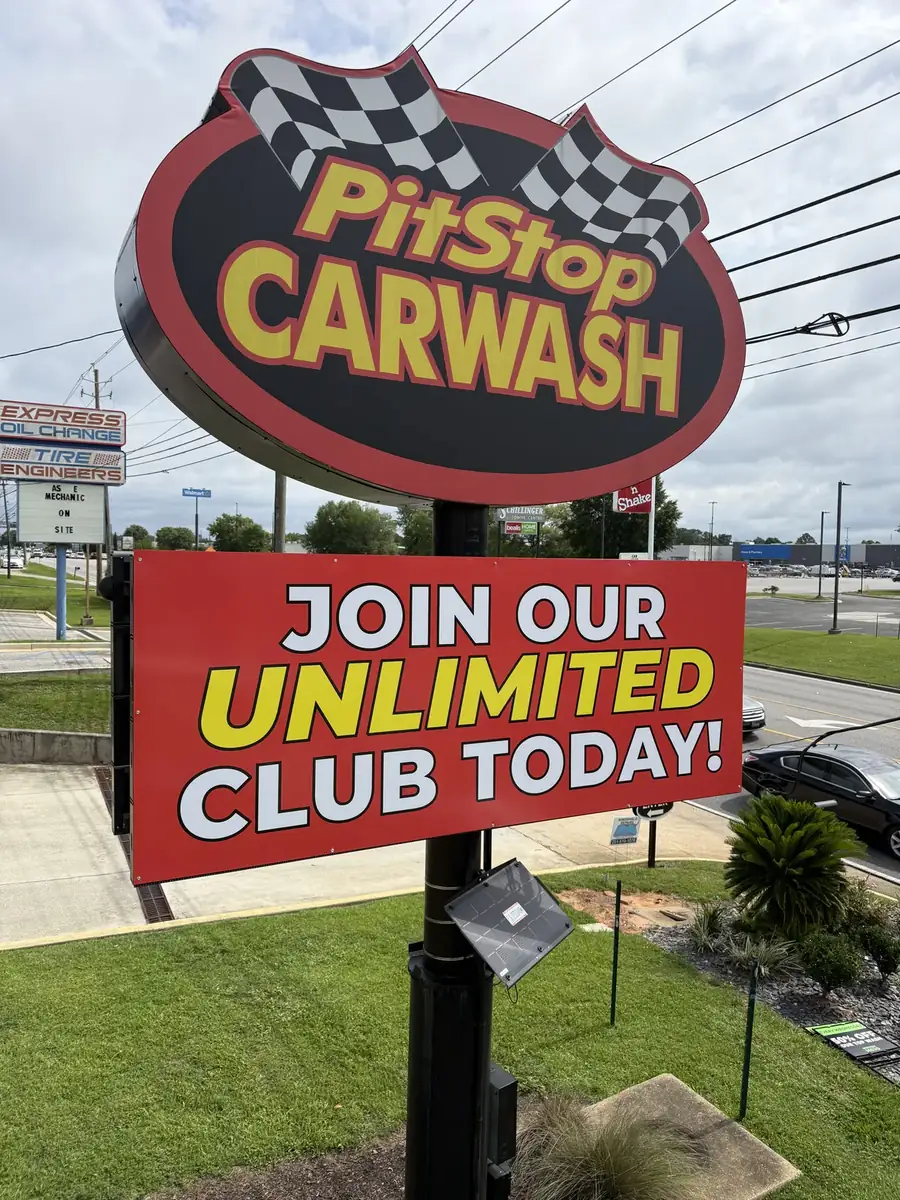 The PitStop Carwash pole sign: a red and black oval logo cabinet with checkered flags above a red Join Our Unlimited Club Today cabinet, on a black pole beside a multi-lane road