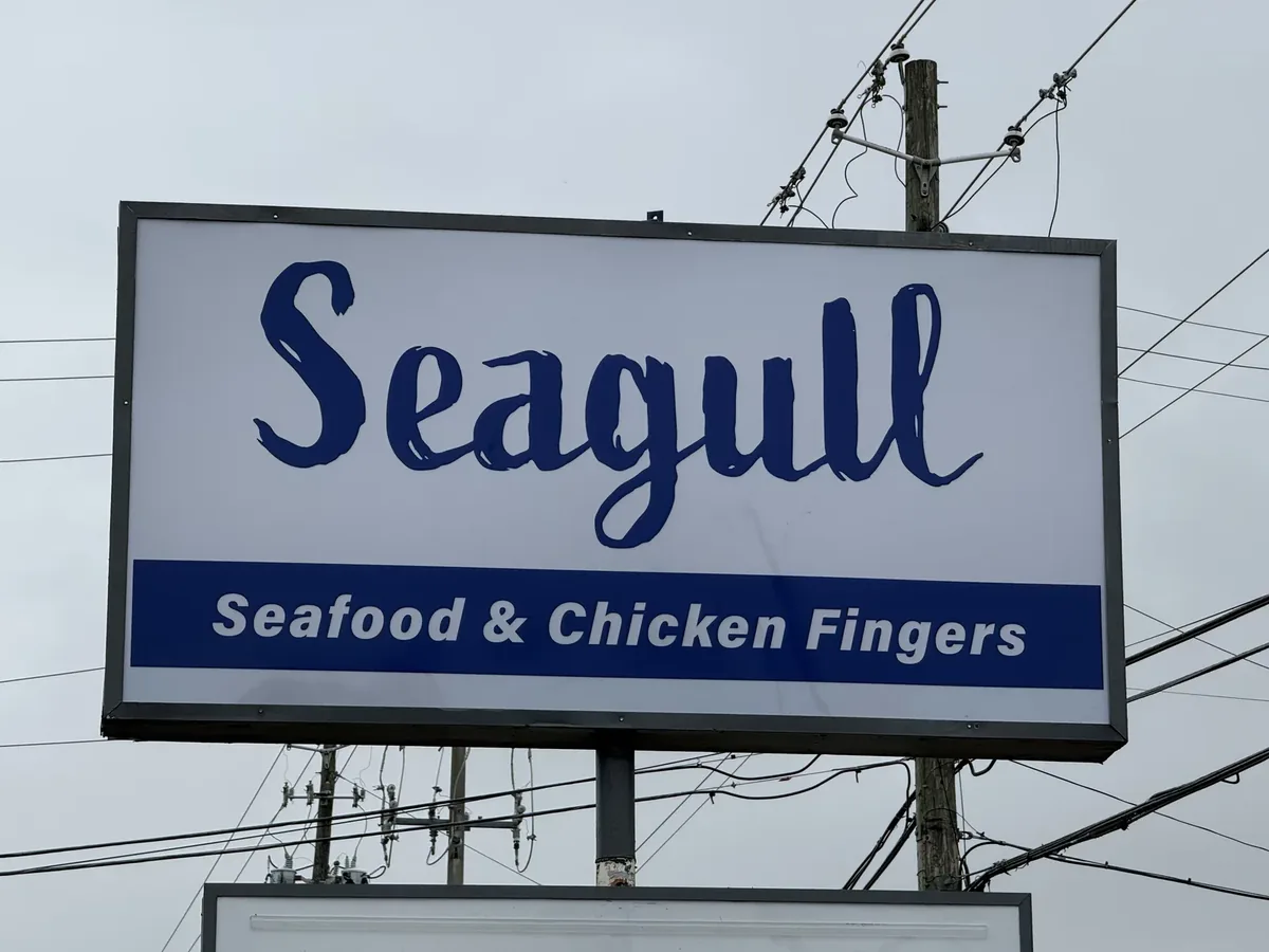 Seagull Seafood & Chicken Fingers sign cabinet on its pole, navy script on a white face against a grey sky