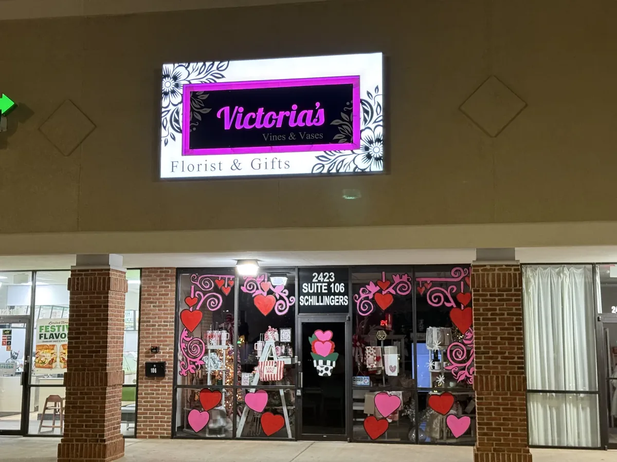 Victoria's Vines & Vases Florist & Gifts sign lit at dusk over a storefront with heart window graphics
