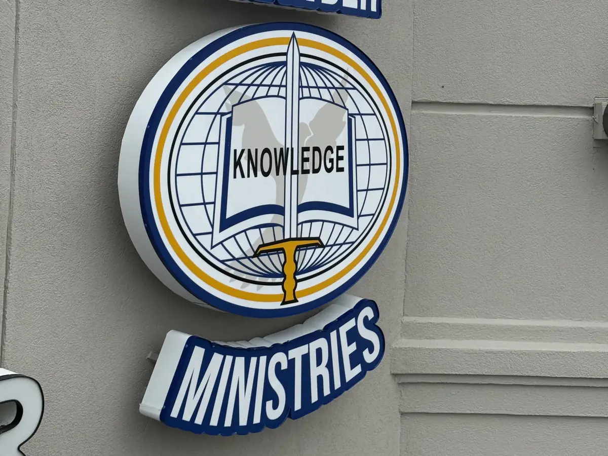 A dimensional round emblem with an open book, a sword and a globe, above a raised MINISTRIES nameplate, mounted on a building's front wall