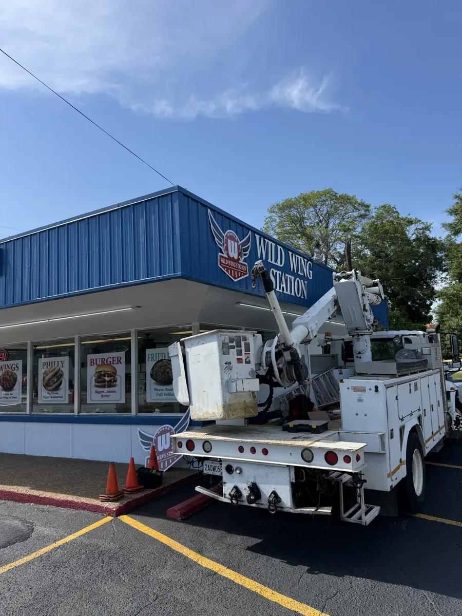 A bucket truck with its empty bucket lowered in front of the Wild Wing Station logo and letters on a blue metal fascia