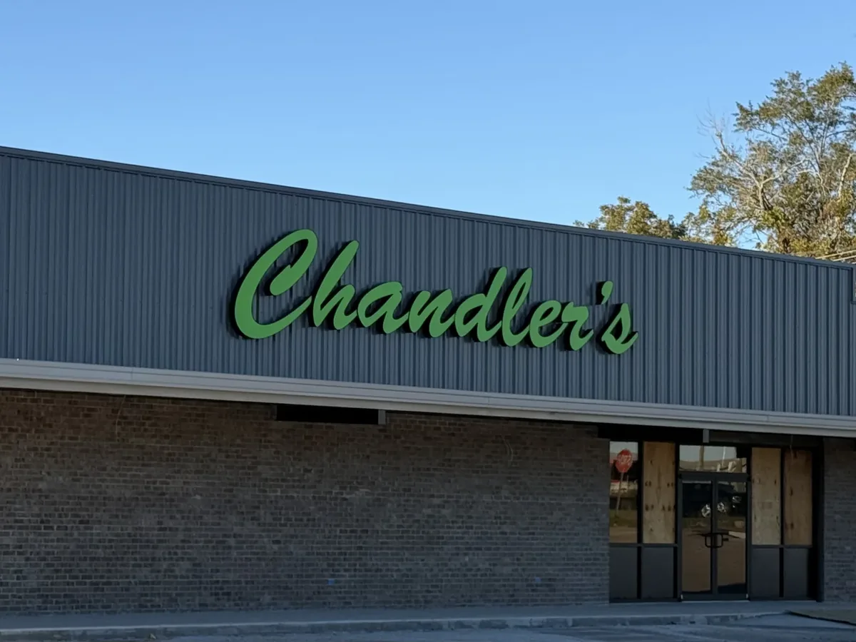 Green script Chandler's channel letters on the grey metal siding of a long brick-based building
