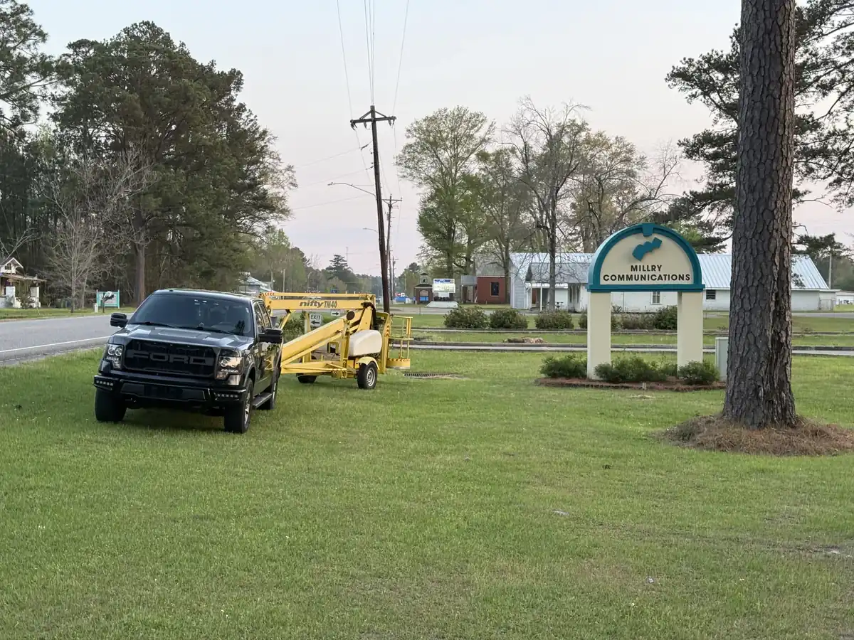 A pickup truck and a yellow towable lift parked on the lawn beside a monument sign at dusk