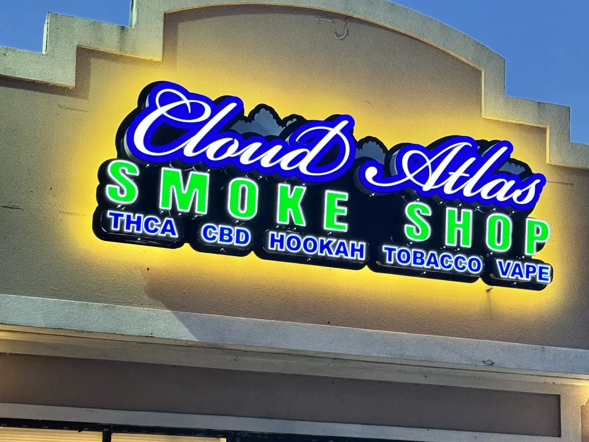 The Cloud Atlas Smoke Shop channel letters lit at dusk, blue script and green block letters with a yellow halo on the storefront wall