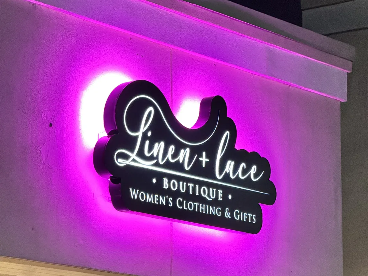 The Linen + Lace Boutique sign lit at night, its shaped black face and script letters glowing with a purple halo on the storefront wall