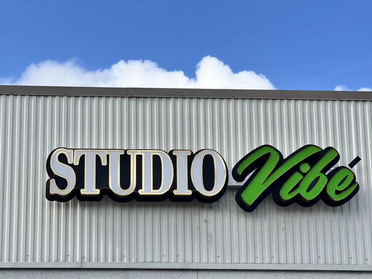 The Studio Vibe channel letters, white block letters and a green script word, on a corrugated metal facade under a blue sky