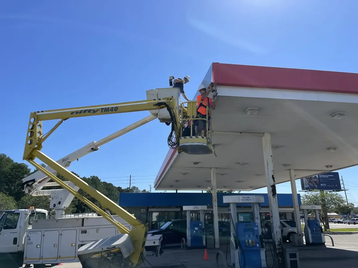 Sign installation crew at a fuel canopy: one installer in an orange high-vis shirt and harness in a towable boom lift, another in the bucket of a bucket truck parked beside it