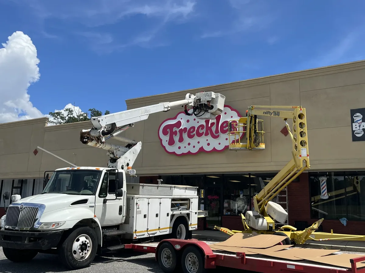 Sign installation at the Freckles storefront: a bucket truck towing a red trailer, with its bucket and a towable boom lift raised at the new sign