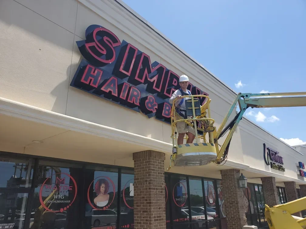 Sign installer standing in a towable lift basket beside large red channel letters on a shopping-center fascia