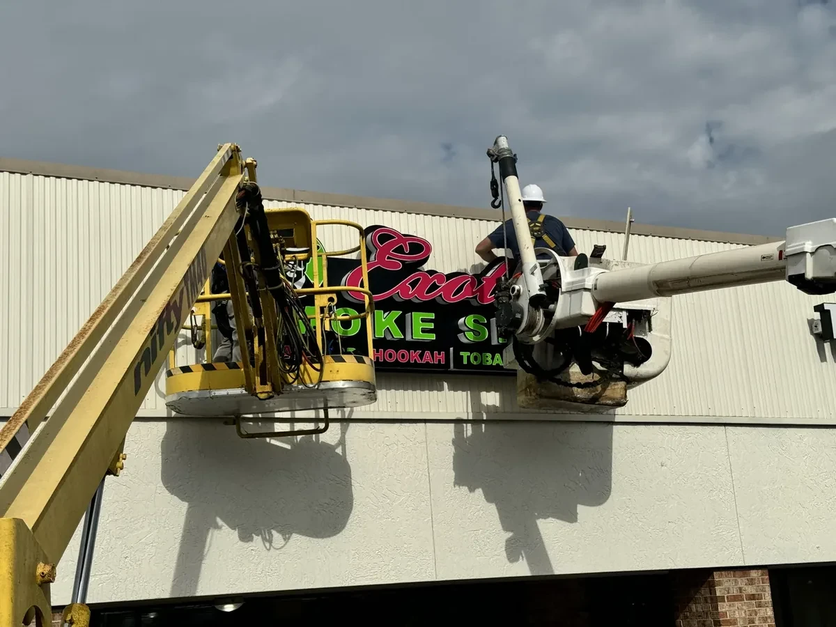 Two installers, one in a bucket truck bucket and one in a towable boom lift, setting the Exotic Smoke Shop sign on a strip-center fascia