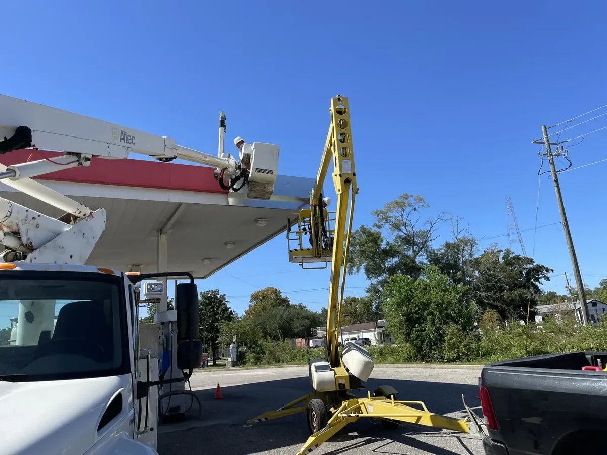 A bucket truck and a towable lift trailer both raised to a fuel-canopy fascia, with a crew member at the top of each