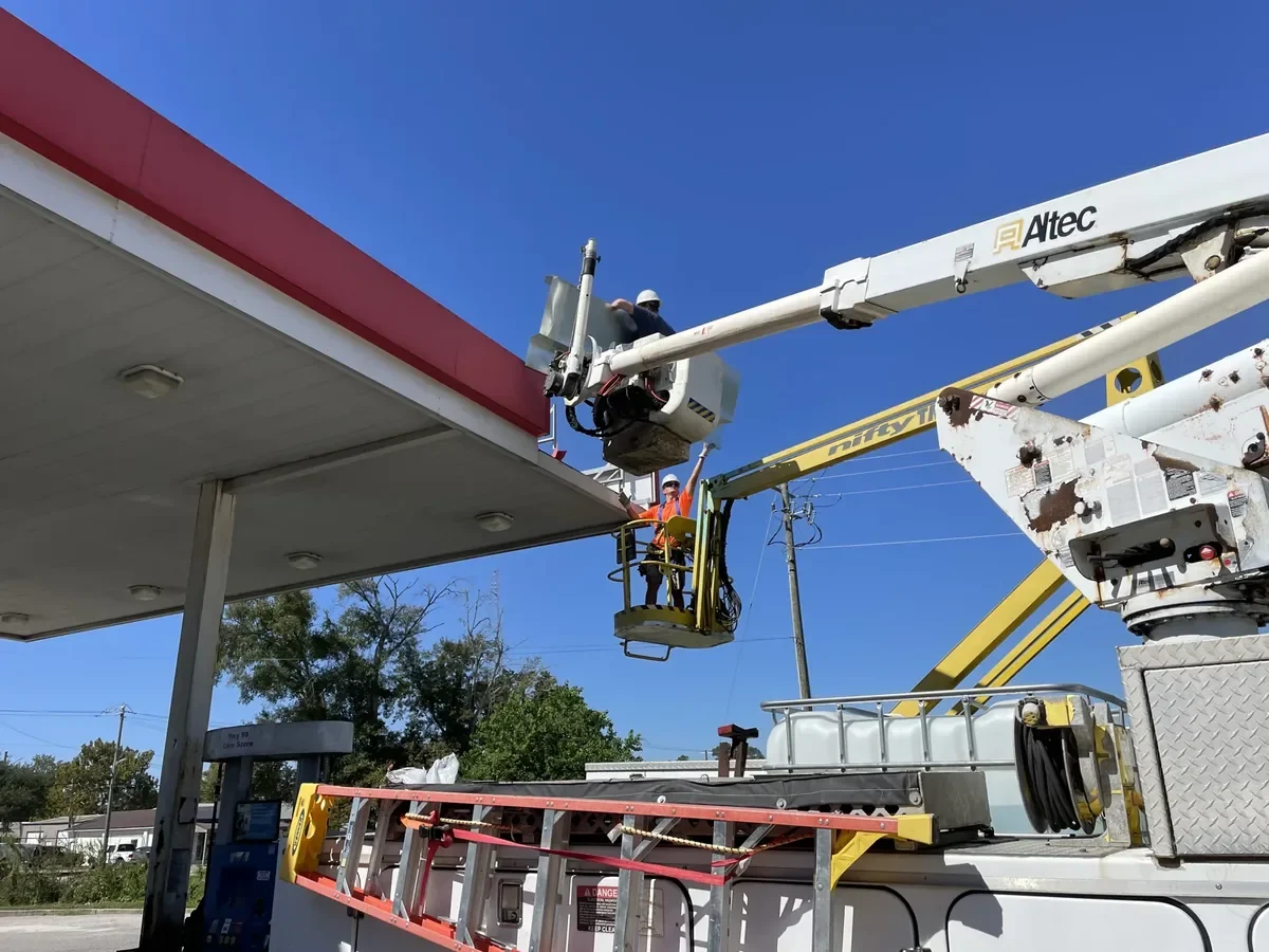 Sparks sign installers at a fuel-canopy fascia, one in a bucket truck raised to the canopy edge and one in a towable lift below it