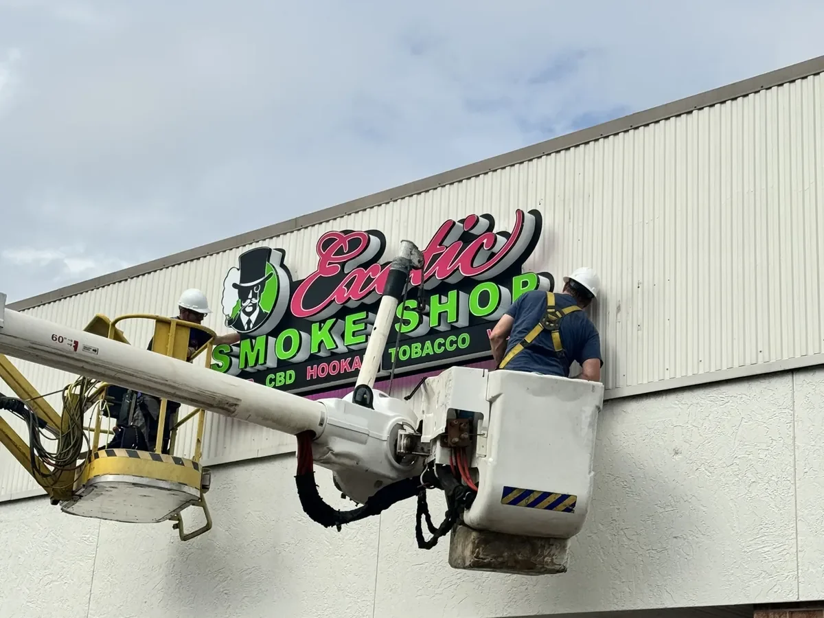 Two Sparks crew members at height in a towable lift and a bucket truck, setting the Exotic Smoke Shop sign on a strip-center fascia
