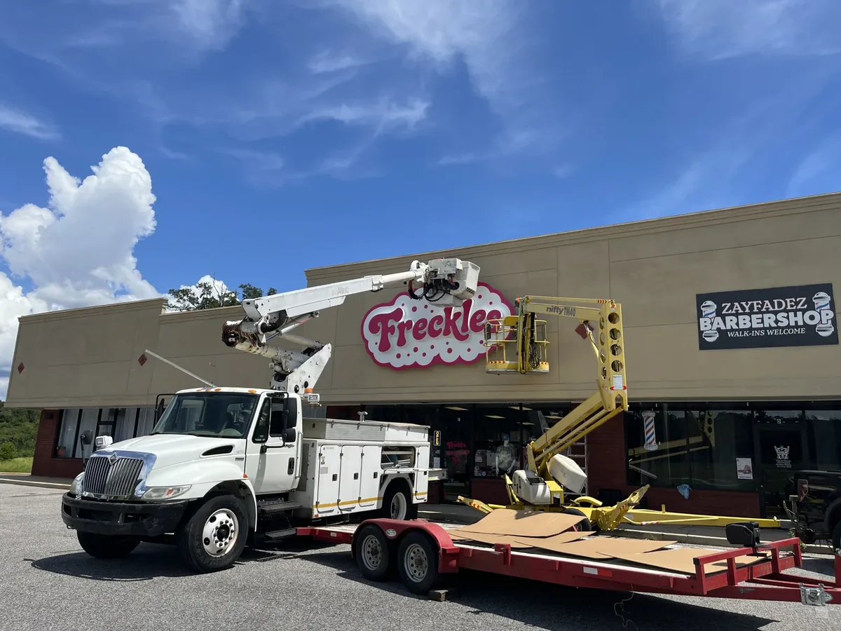 A Sparks bucket truck with its boom raised at the Freckles storefront sign, and a towable lift on its trailer