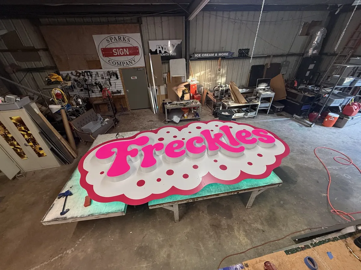 The pink Freckles sign laid on tables on the shop floor, with the oval Sparks Sign Company logo on the wall behind