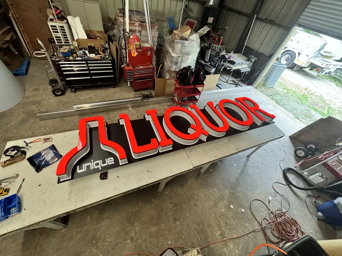 Red Unique Liquor channel letters on a shop bench, with tool chests behind and a bucket truck outside the open shop door