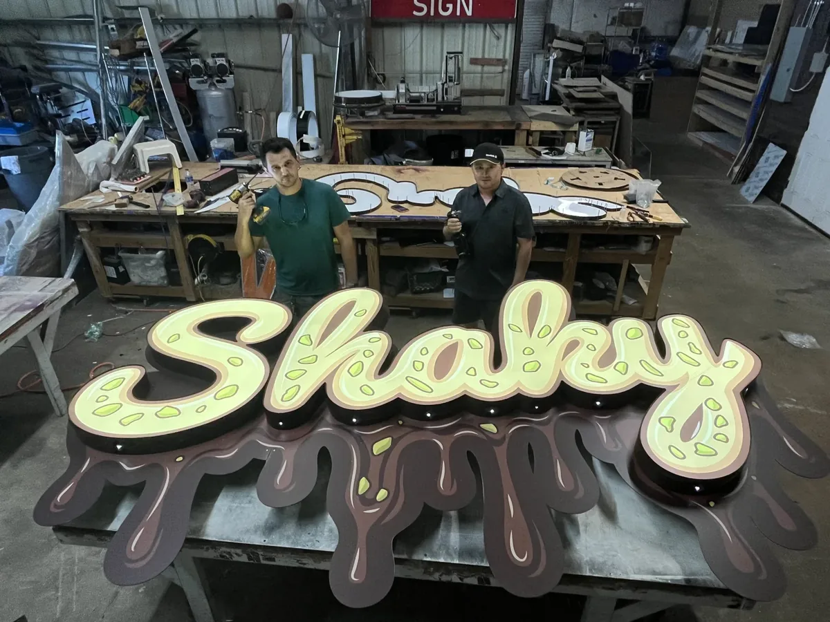 Two Sparks crew members holding drills behind a lit script channel-letter sign on a chocolate-drip backer in the sign shop in Mobile, Alabama