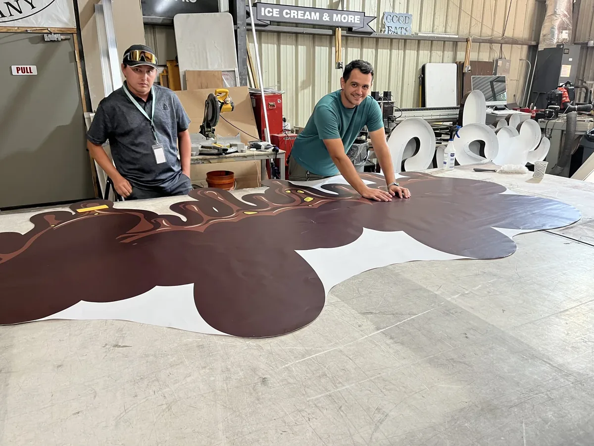 Two Sparks crew members smoothing printed vinyl onto a large shaped sign backer laid across the shop floor