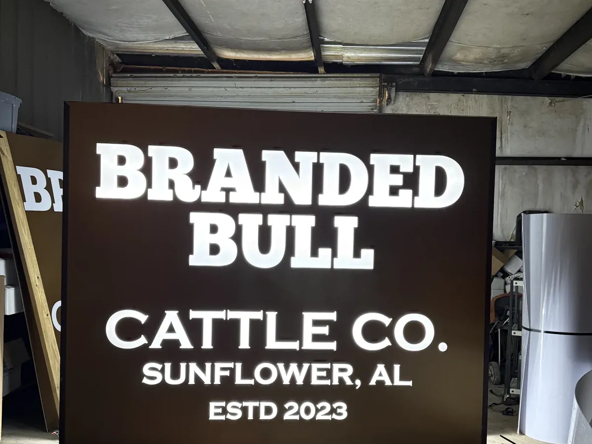 A lit cabinet sign (light box) standing in the sign shop, white letters glowing on a dark brown face