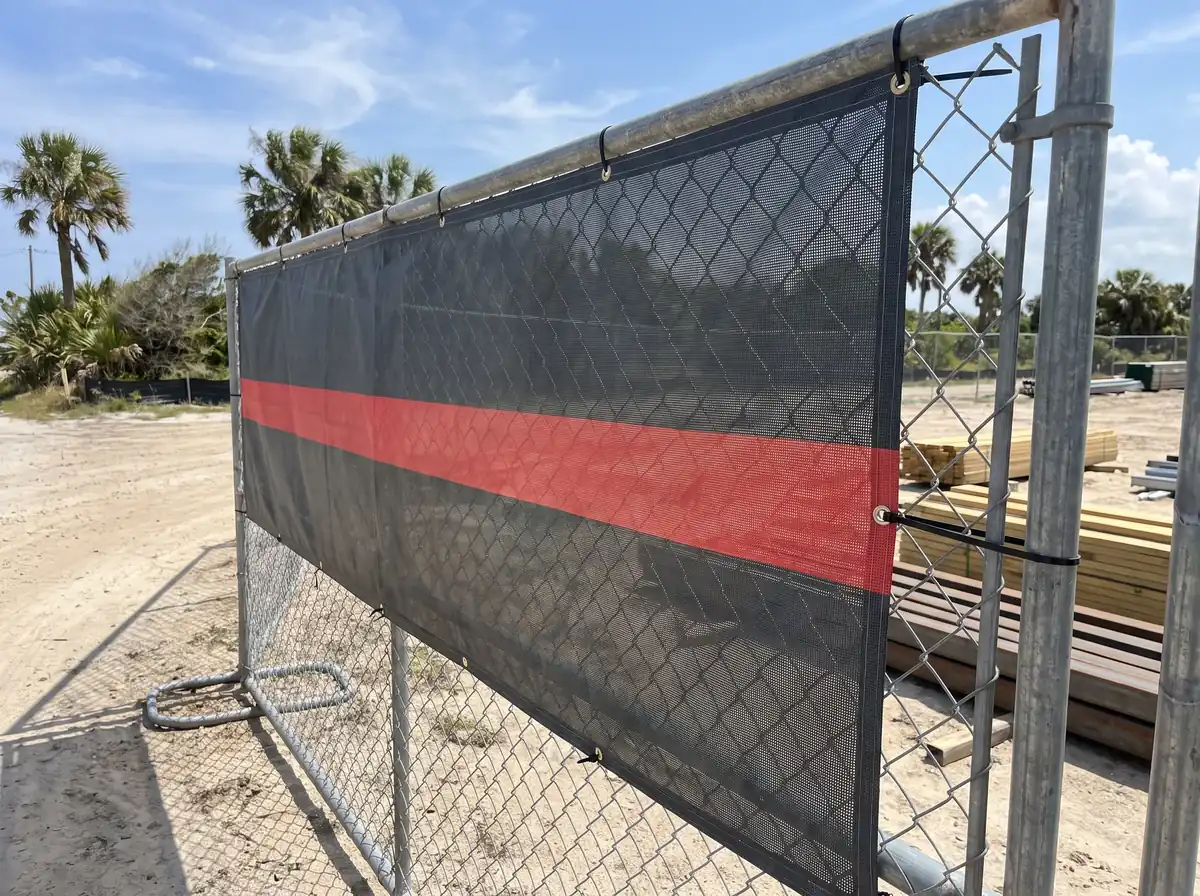 Charcoal mesh banner with a red stripe zip-tied through its grommets along a chain-link fence, light showing through the mesh